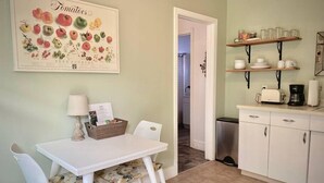 Une cuisine avec une table blanche, des chaises et une estampe botanique encadrée. On y trouve des étagères remplies de vaisselle et une machine à café.
