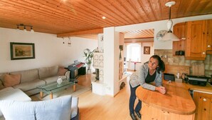 A modern living room with a wooden ceiling, a glass coffee table, a sofa, and a kitchen area with wooden cabinets and a sink. - Upper mill (Sohland an der Spree)