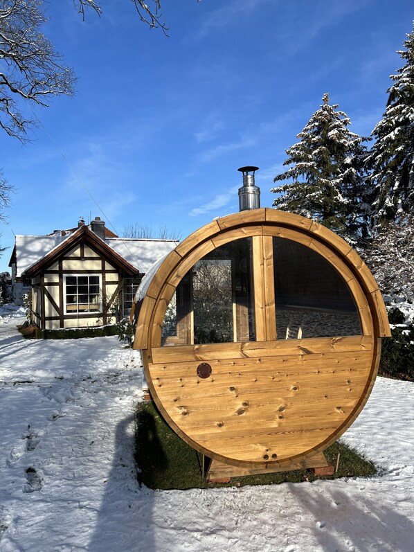 A wooden barrel structure with a chimney, set in a snowy landscape with a house in the background.