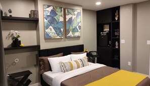 Quarto com cama, mesa de cabeceira, prateleira com objetos decorativos e duas pinturas abstratas na parede.