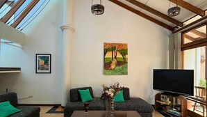 A spacious living room with a high ceiling, wooden beams, a large TV, and a painting on the wall. - Gaudys Hotel (San José)
