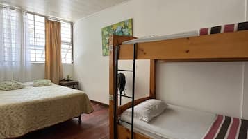 Una habitación con una cama individual, una litera, suelo de madera, ventilador de techo y una ventana con cortinas.