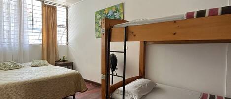 A room with a bunk bed, a single bed, wooden flooring, a ceiling fan, and a window with curtains.