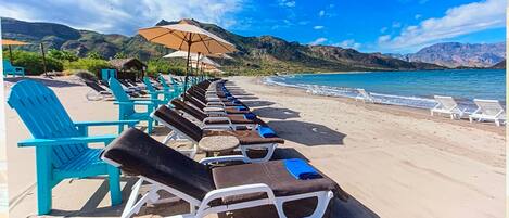 A beach with rows of sun loungers and umbrellas, a clear sky, and mountains in the background.