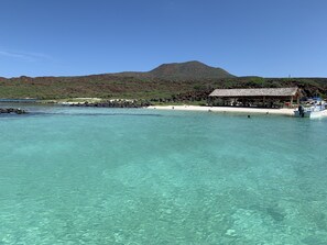 En strand med klart turkis vand, en hytte med stråtækt tag og et bjerg i baggrunden.