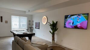 Ein modernes Wohnzimmer mit Poolbillard, einer bequemen Couch, einem Couchtisch, einer Küstpflanze und einem Fernseher, der eine Szene am Pool zeigt.