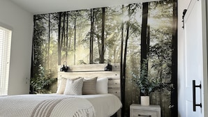 Ein Schlafzimmer mit einer großen Waldmalerei an der Wand, einem Bett mit weißer Bettwäsche, einem Nachttisch und einem Fenster mit Jalousien.