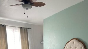 Une chambre à coucher avec un lit, une table de chevet en bois, un ventilateur de plafond et une fenêtre avec des rideaux.