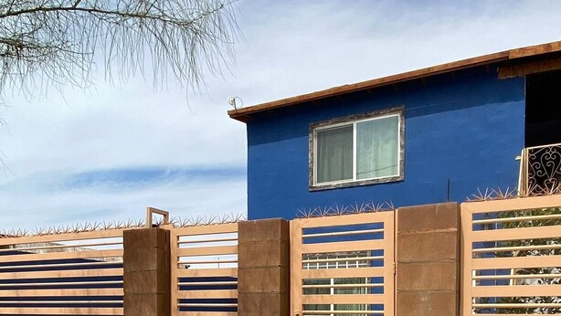 Une entrée fermée par un portail, avec un bâtiment bleu, une fenêtre et un portail en métal.