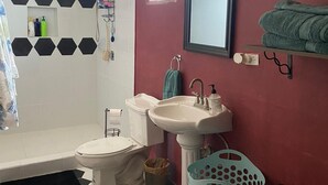Une salle de bain avec des toilettes blanches, un lavabo, un miroir et une douche. On peut voir des serviettes et des articles de toilette.