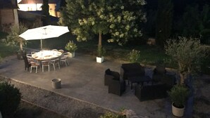 Une terrasse extérieure avec une table ronde et des chaises, entourée de plantes en pot et d’un grand parasol.