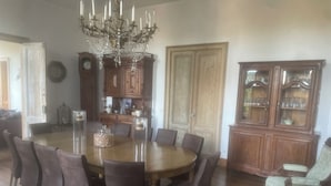 Une salle à manger avec une grande table ronde, des chaises sombres, un lustre, des meubles en bois et une horloge.