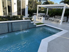 Un área de piscina exterior moderna con una cascada, rodeada por una pérgola blanca y mobiliario.