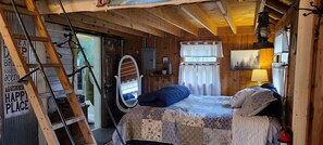 A cozy cabin room with a bed, a ladder, a mirror, and a window with curtains. - Wood Haven Lodge. Secluded, cozy log cabin in Northern Michigan. (South Boardman)