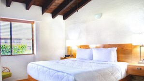 Quarto com cama de madeira, roupa de cama branca, mesa de cabeceira com abajur e janela com persianas.