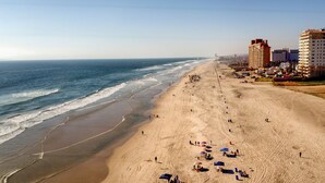 Uma praia com uma fileira de edifícios ao longo da costa, incluindo um hotel com vários andares e varandas.