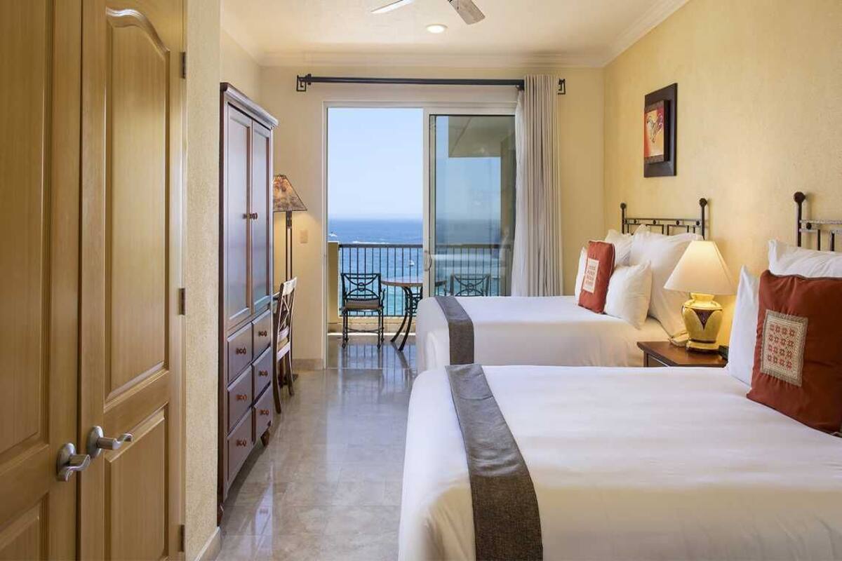 Habitación de hotel con dos camas, un balcón con vistas al océano, un escritorio con lámpara y una silla.