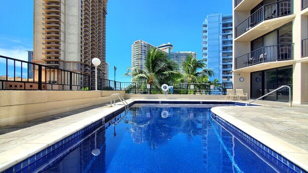 A swimming pool with a tiled edge, surrounded by a low fence, and palm trees in the background. - Large Studio w/ Pool sleeps six in comfort PLUS FREE PARKING INCLUDED! (Honolulu)