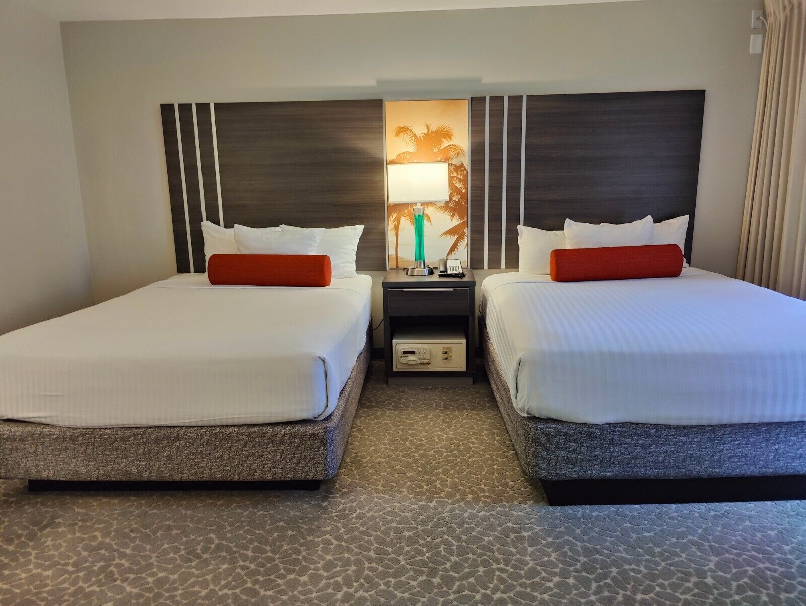 Une chambre d’hôtel avec deux lits, chacun agrémenté de draps blancs et de coussins décoratifs rouges, une table de chevet avec une lampe et une moquette à motifs.