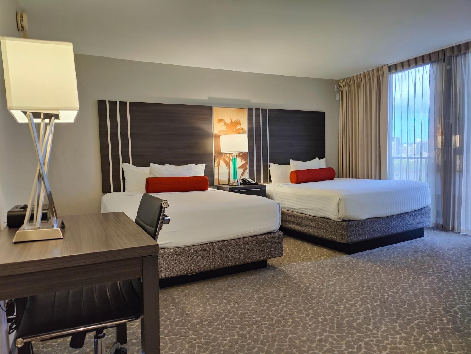 Two queen beds with red bolster pillows sit against dark wood headboards illuminated by a palm tree lamp.