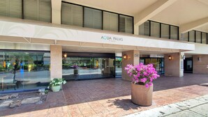 The exterior of Aqua Palms at Waikiki, featuring a modern entrance with glass doors, a stone pillar, and a large planter with pink flowers. - Large Studio w/ Pool sleeps six in comfort PLUS FREE PARKING INCLUDED! (Honolulu)