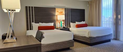 A hotel room with two beds, a desk, a lamp, and a view of the city.