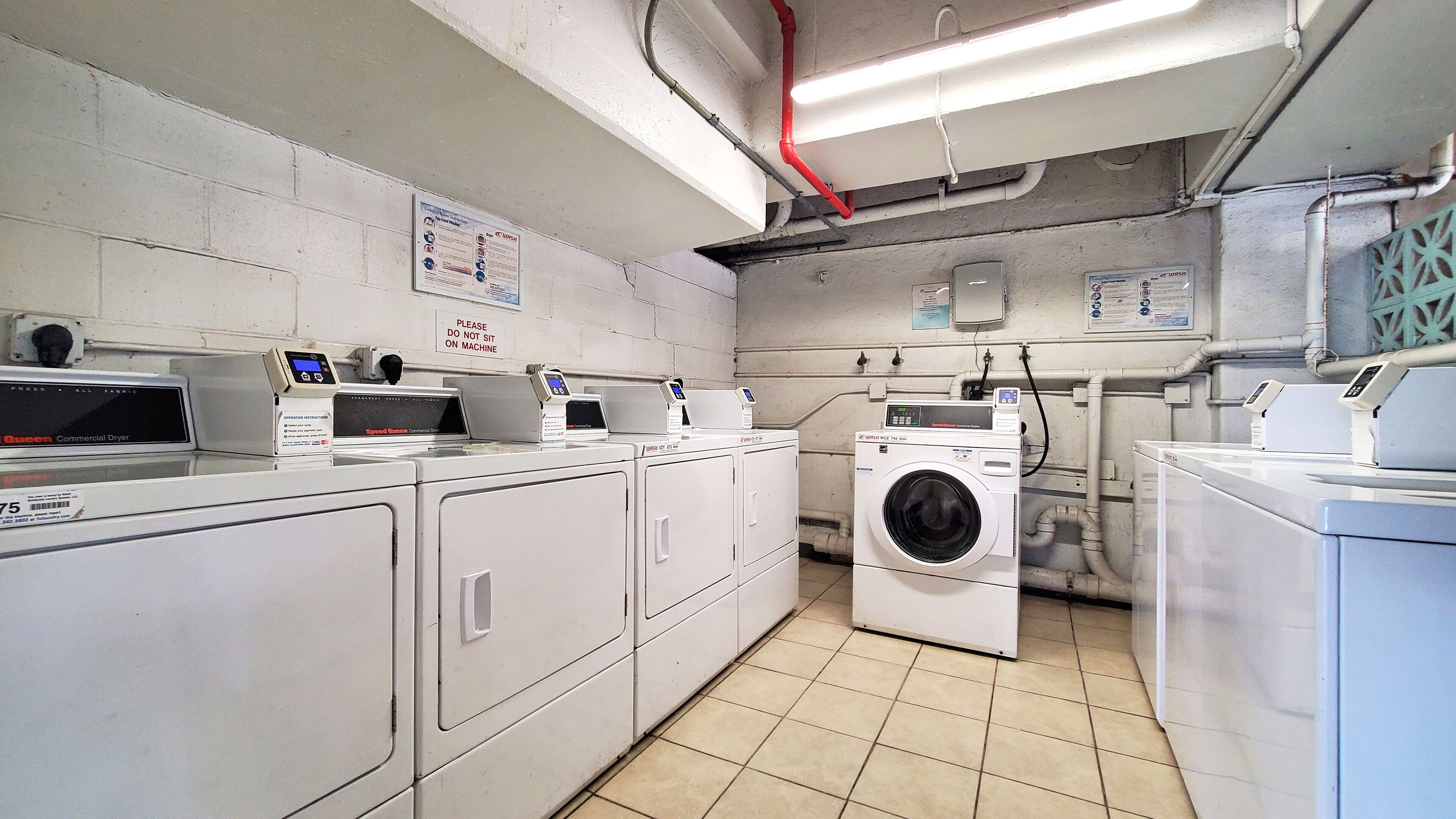 A laundry room with commercial dryers and washing machines, a warning sign on the wall, and visible plumbing.