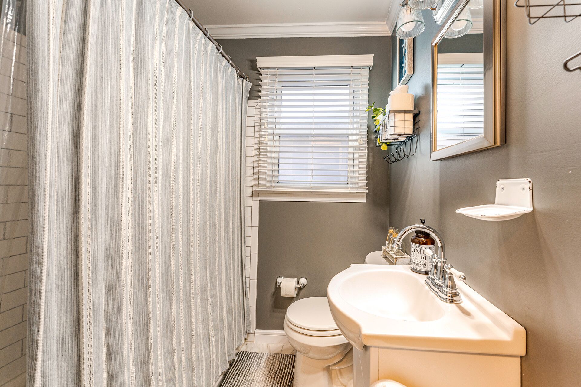 Ein Badezimmer mit Duschvorhang, Toilette, Waschbecken mit Seifenspender und einem Fenster mit Jalousien.