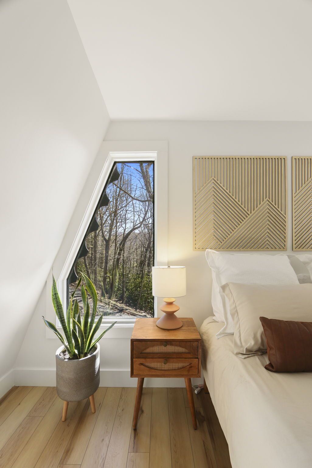 A bedroom with a skylight, a bedside table with a lamp, a potted plant, and a bed with pillows.
