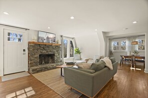A spacious living room with a stone fireplace, a grey sofa, wooden flooring, and a dining area with a table and chairs. - A-Frame in the heart of Boone (Boone)