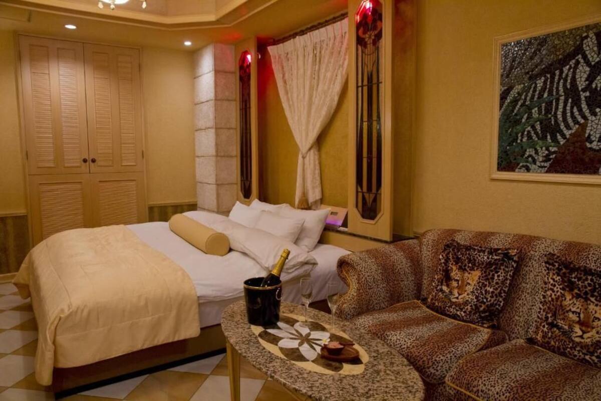 A hotel room with a bed, a sofa, a small table with a bottle and glass, and a painting on the wall.