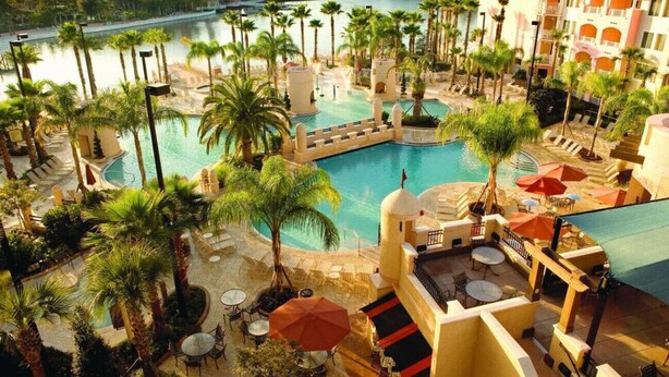 Ett semesterhotell med en stor pool i mitten, omgivet av palmer och utomhusområden med sittplatser.