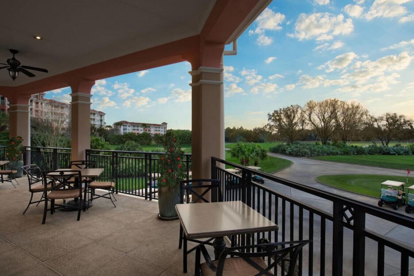 A covered outdoor seating area with tables and chairs, overlooking a golf course and a building in the background.