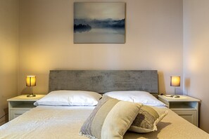 Un dormitorio ordenado con una cama grande, dos mesitas de noche con lámparas y una obra de arte colgada en la pared.