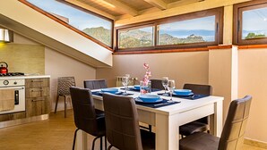 A dining area with a table set for four, a kitchen with a stove, and large windows offering a view of the hills. - Villetta Desiderio Apartment (Giardini Naxos)