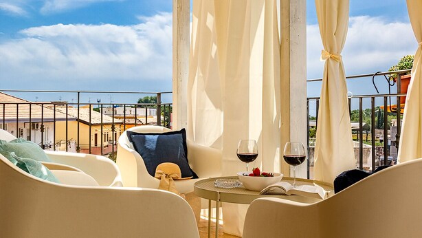 A balcony with white chairs, a table with a wine glass and strawberries, and a view of buildings and sky. - Villetta Desiderio Apartment (Giardini Naxos)
