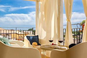 Ein Balkon mit weißen Stühlen, einem Tisch mit Weinglas und Erdbeeren sowie Blick auf Gebäude und Himmel.