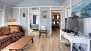 A room with a wooden floor, a brown leather sofa, a wooden coffee table, a TV on a white stand, and a painting on the wall. - Bahama Waves (Jamaica Beach)