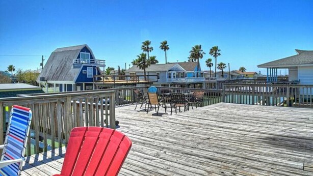 A wooden deck with chairs and tables, overlooking a beach area with palm trees and a blue building. - Bahama Waves (Jamaica Beach)