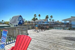 A wooden deck with chairs and tables, overlooking a beach area with palm trees and a blue building.