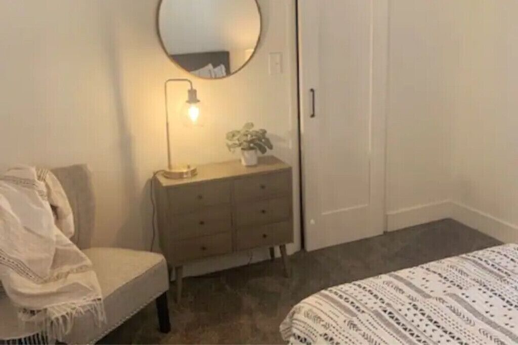 A bedroom with a bed, a dresser, a chair, a mirror, and a bedside lamp.