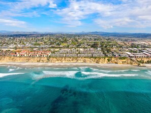 An aerial view of a coastal town with beachfront hotels, clear blue waters, and a hilly landscape in the background.
