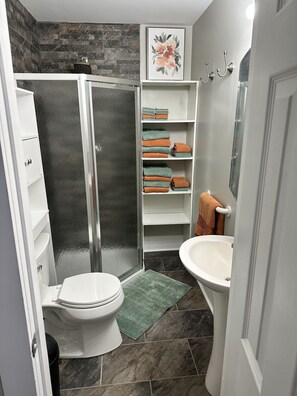 A bathroom with a glass-enclosed shower, a toilet, a sink, and shelves with folded towels.