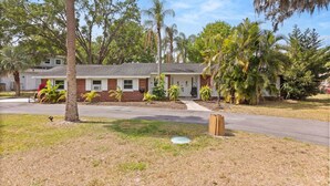 A single-story house with a brick facade, a front porch, and a lawn with various plants and trees.