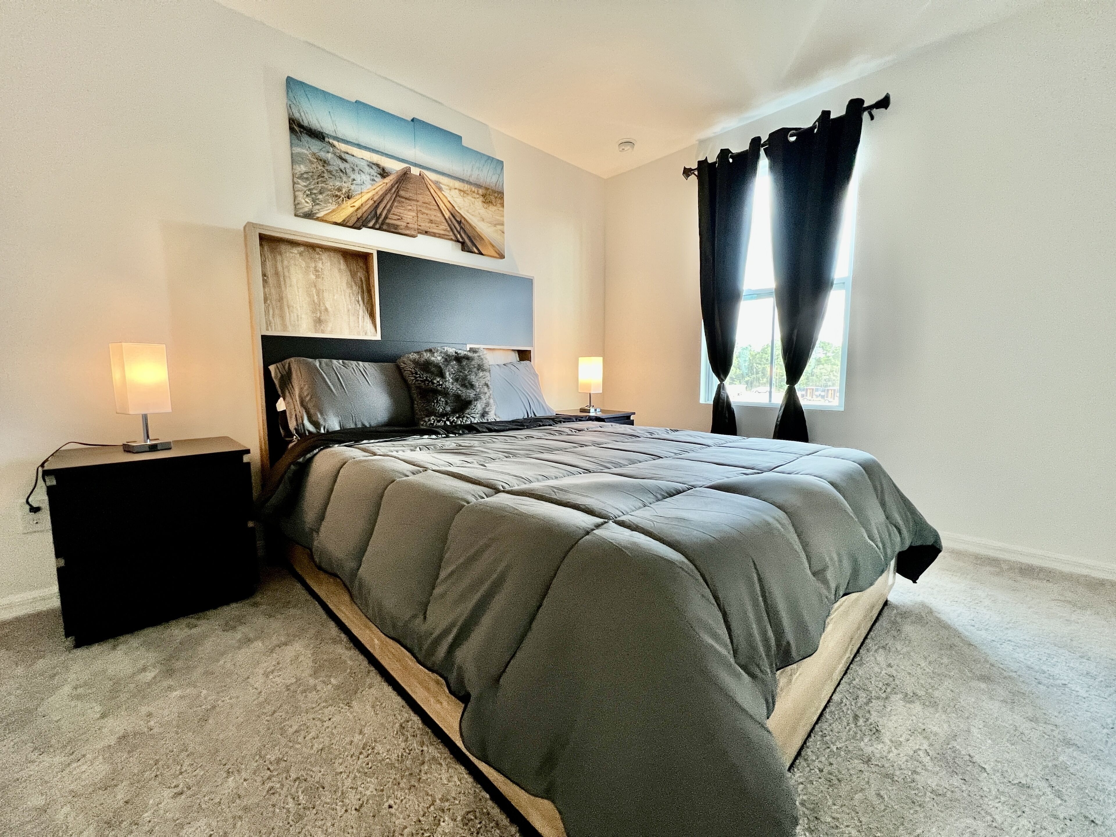 A bedroom with a large bed, two bedside tables, a nightstand, a painting above the bed, and a window with curtains.