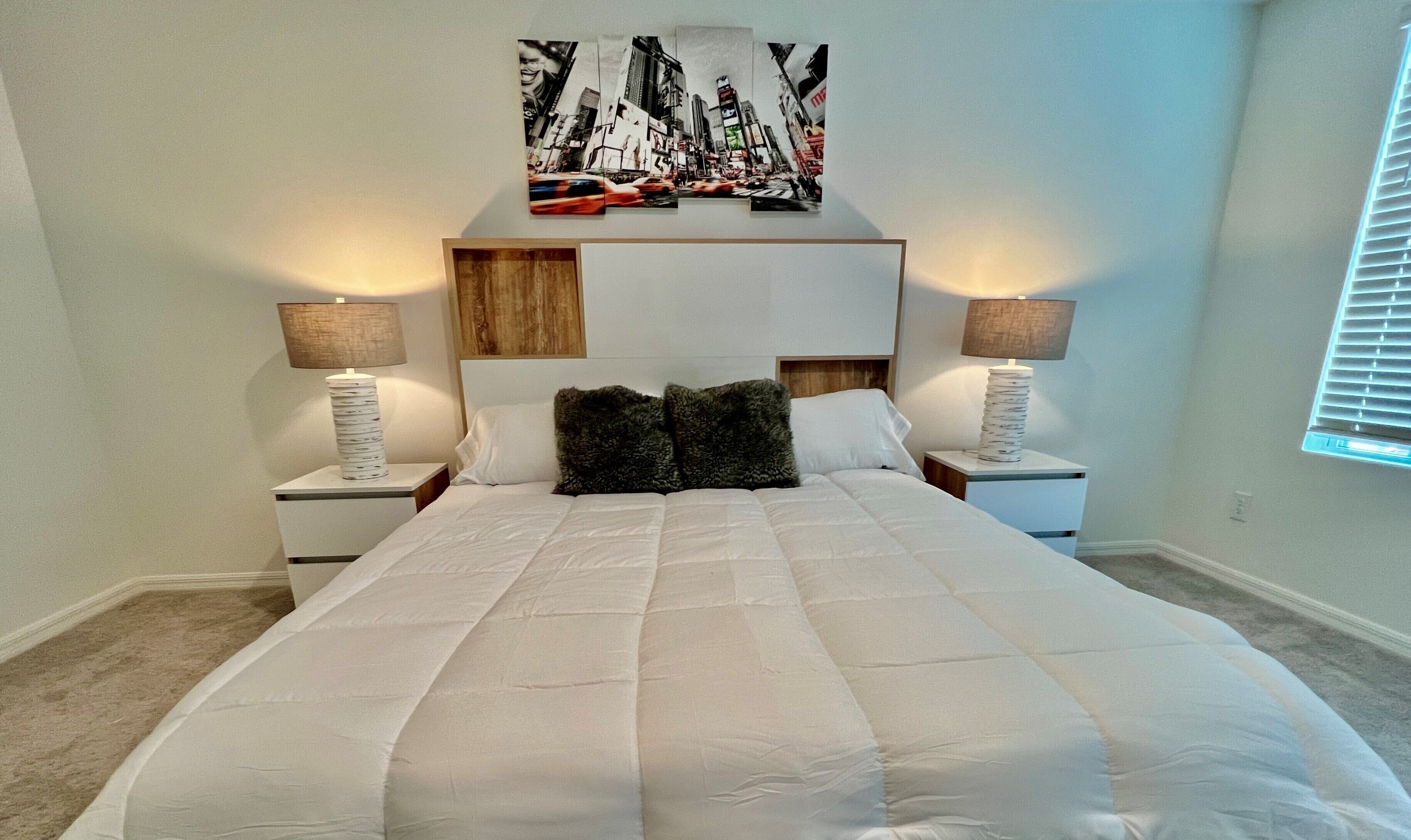 A bedroom with a large bed, two bedside tables with lamps, a headboard with a cityscape artwork, and a window with blinds.