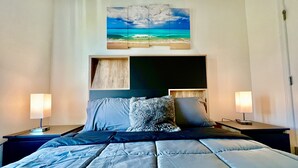 Un dormitorio con una cama, dos mesitas de noche, una mesita auxiliar, un cabecero y un cuadro con una escena de playa.