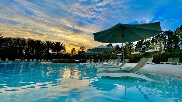 Una piscina con sillas de descanso y sombrillas, rodeada de árboles y un cielo al atardecer.