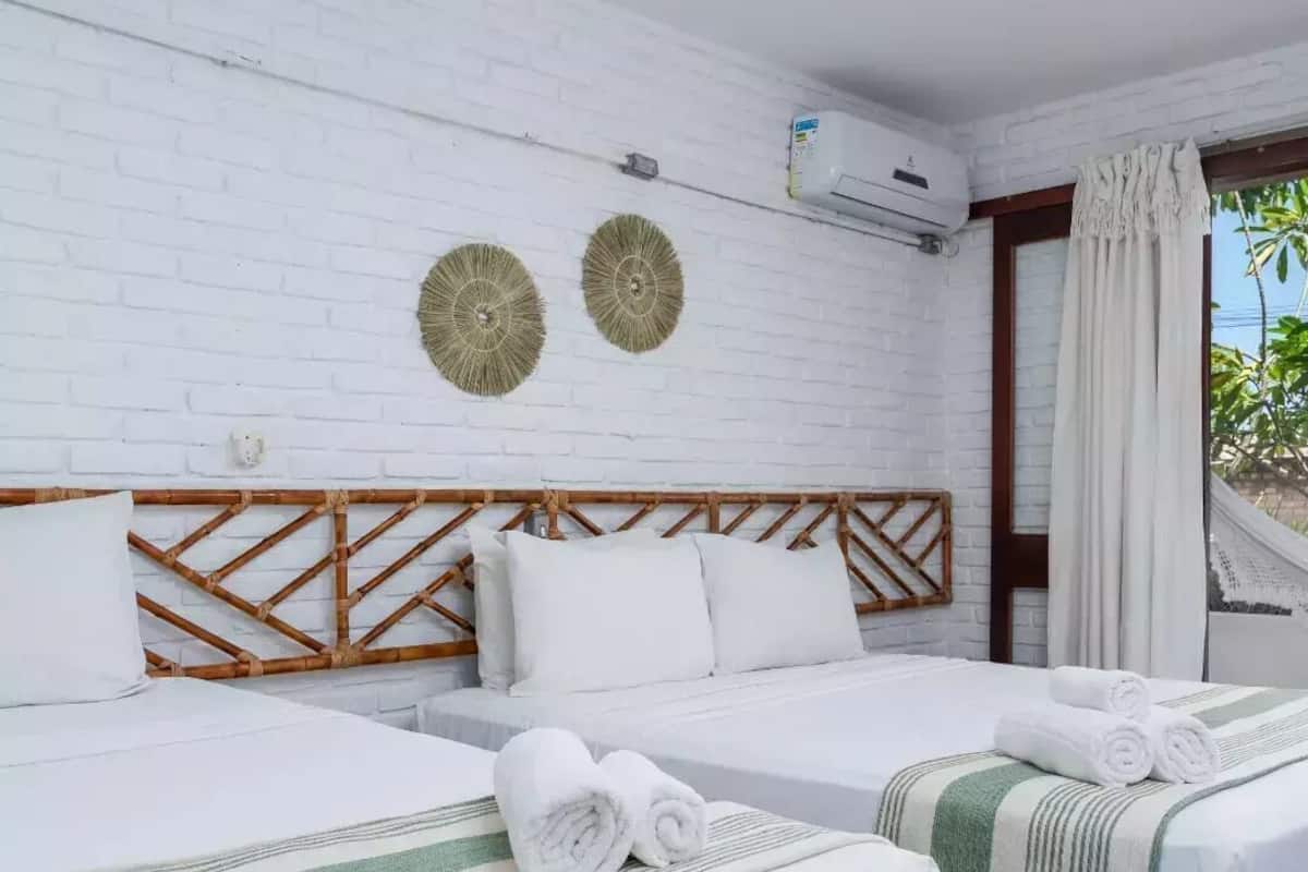 Ein Hotelzimmer mit zwei Betten, weißer Bettwäsche, einer weißen Ziegelwand, einer Klimaanlage und Blick ins Grüne.