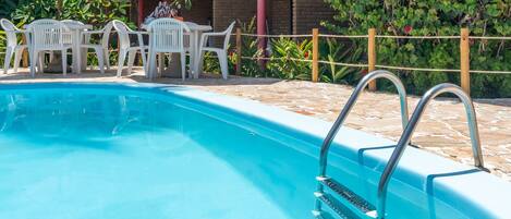 A swimming pool with a tiled deck, surrounded by white plastic chairs and umbrellas.